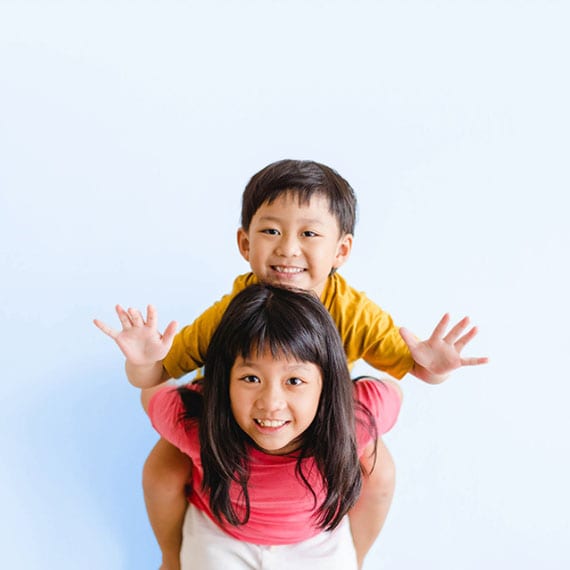 A young child in a yellow shirt sits on the shoulders of a student in a pink top against a plain light blue background.