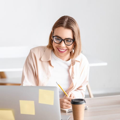 A teacher at Connections Academy in a beige button down shirt writhing something down on a piece of paper while smiling at her laptop.