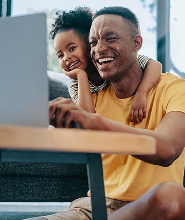 A father being hugged by his daughter from behind while looking at a laptop screen.