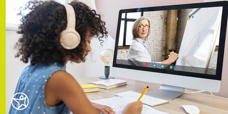 A high school student wearing a blue shirt watches an online lesson on a monitor while wearing headphones.